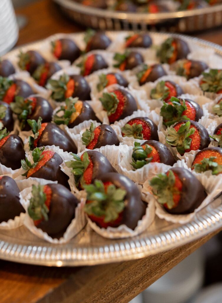 A large silver tray filled with chocolate-covered strawberries, each placed in a white paper cup, displayed on a wooden surface at a Chastain Horse Park wedding reception.
