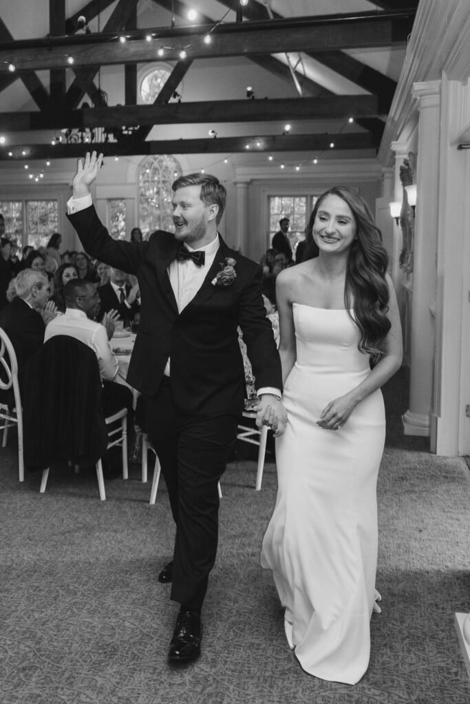 A newlywed couple enters their Chastain Horse Park wedding reception, holding hands and smiling; the groom waves enthusiastically while guests seated at tables watch and celebrate beneath twinkling lights and wooden beams overhead.