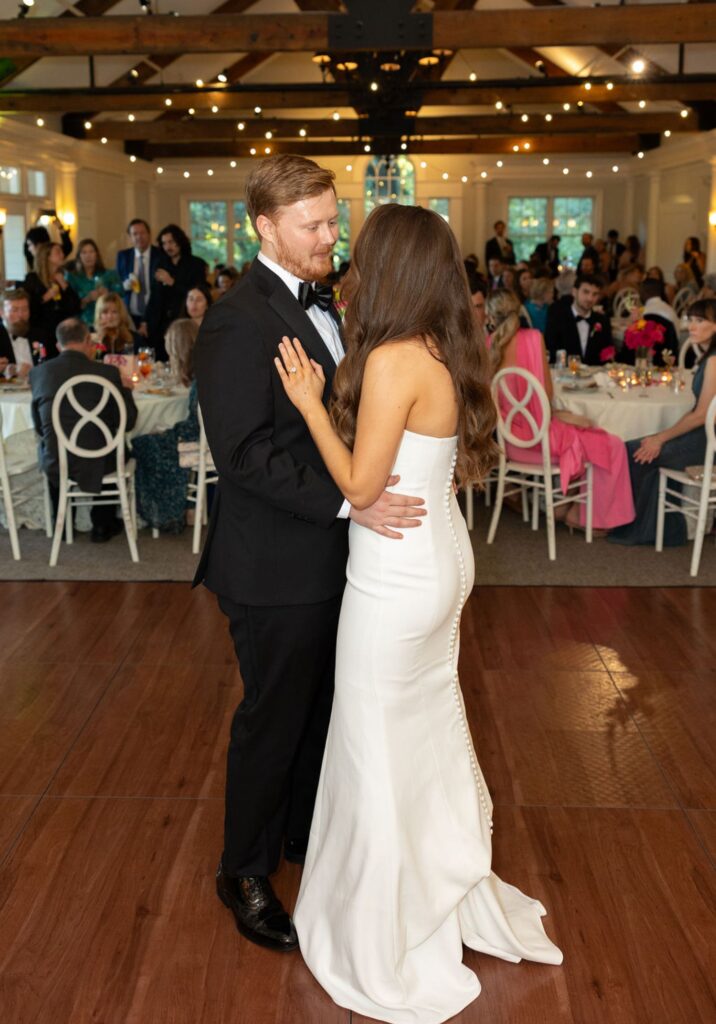 A bride in a white strapless gown and a groom in a black tuxedo share their first dance at their Chastain Horse Park wedding reception, surrounded by seated guests and warm string lights in a beautifully decorated hall.