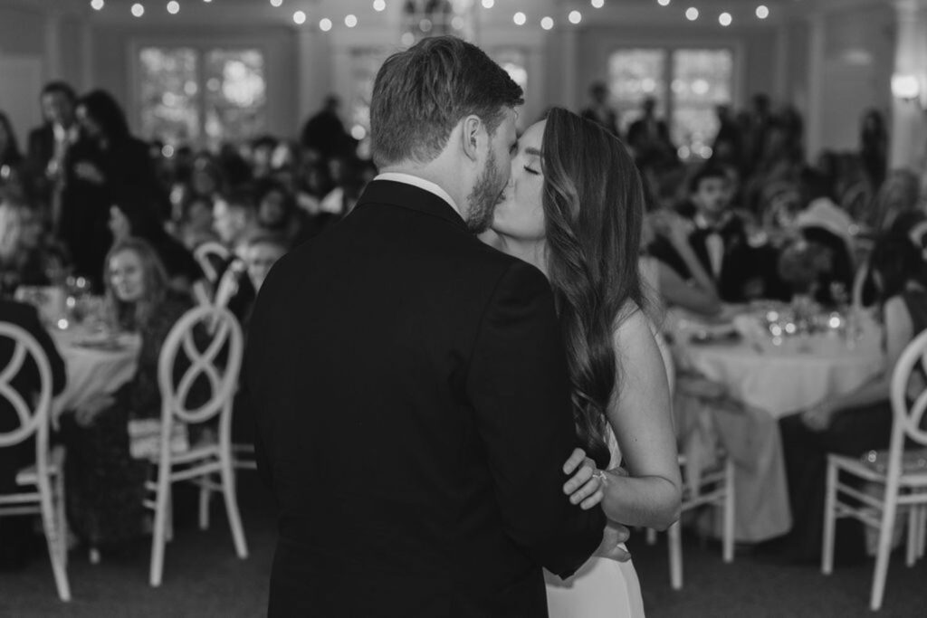 A bride and groom share a kiss while dancing at their Chastain Horse Park wedding reception, surrounded by seated guests in a decorated, warmly lit venue.