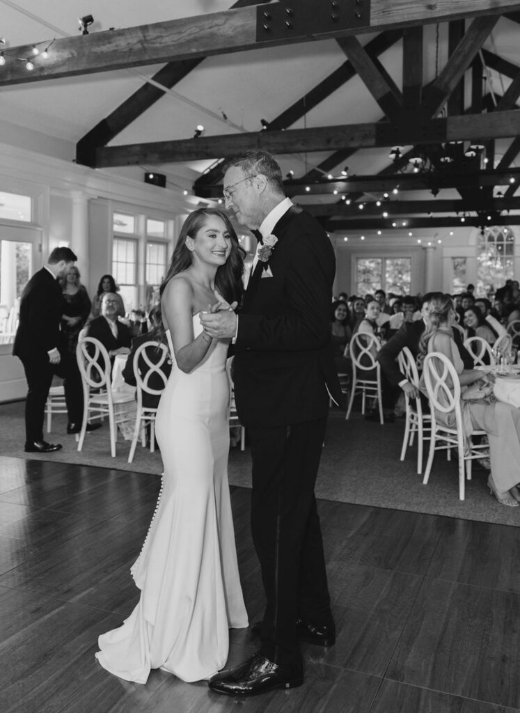 A bride and groom share a dance on a wooden floor in a decorated Chastain Horse Park wedding reception hall, surrounded by smiling guests. The elegant, joyful scene is beautifully captured in black and white.