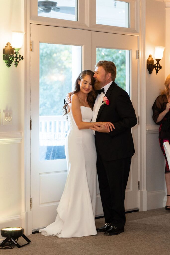 A bride in a white dress and a man in a black suit share a tender dance, smiling and embracing near glass doors in a softly lit room during their Chastain Horse Park wedding reception.