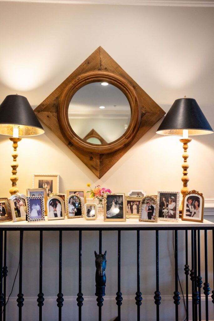 A collection of framed family photos from a Chastain Horse Park wedding reception sits on a table beneath a wooden-framed round mirror and two lit lamps with black shades, against a cream-colored wall.