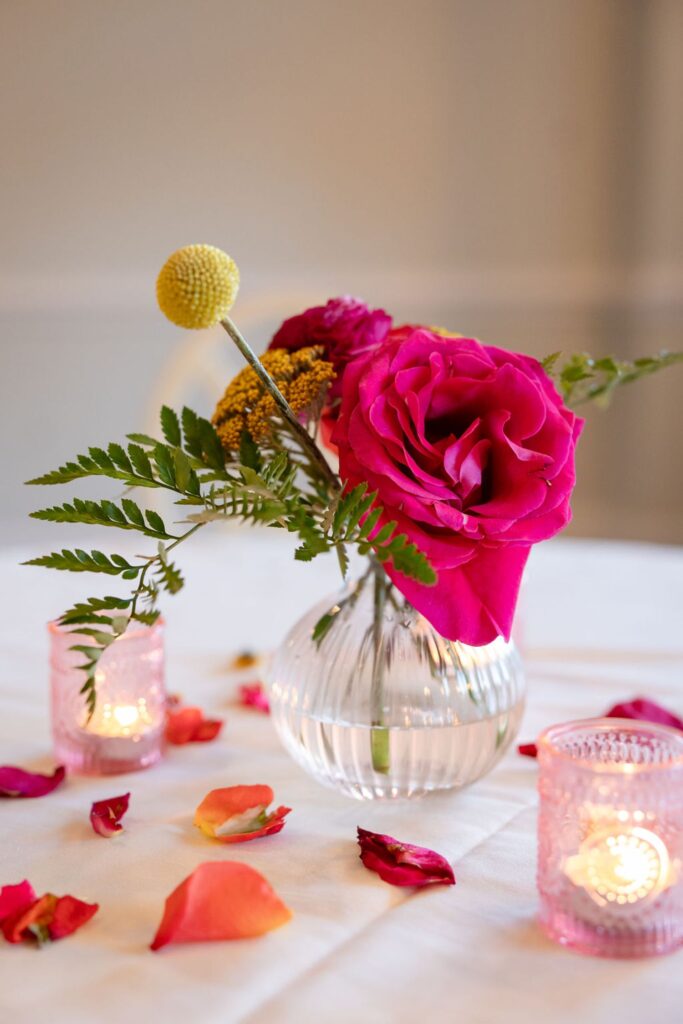 At a Chastain Horse Park wedding reception, a small glass vase holds bright pink roses, yellow flowers, and greenery on a white tablecloth, surrounded by scattered rose petals and two lit pink votive candles.