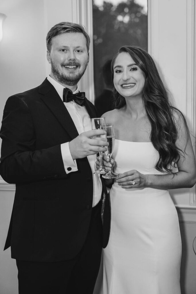 A smiling couple dressed formally, the man in a black tuxedo and the woman in a white dress, hold champagne glasses and stand close together at their Chastain Horse Park wedding reception in front of a window.