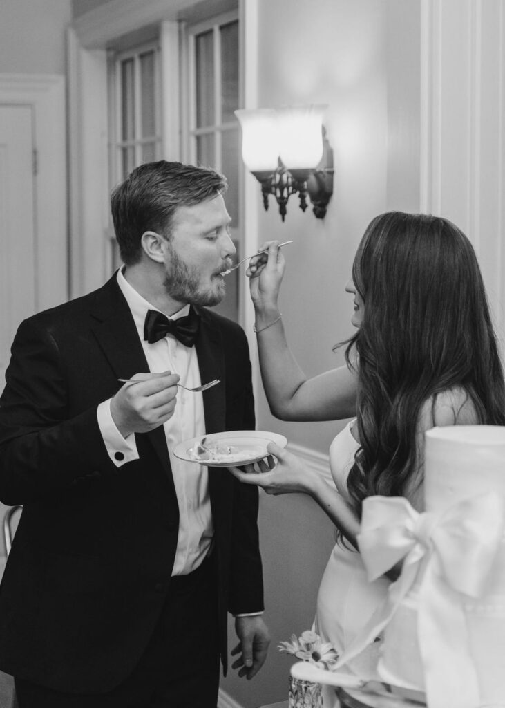 A bride and groom feed each other cake at their Chastain Horse Park wedding reception, smiling and standing close together. The groom wears a tuxedo and the bride wears a strapless dress, lit by soft indoor lighting.