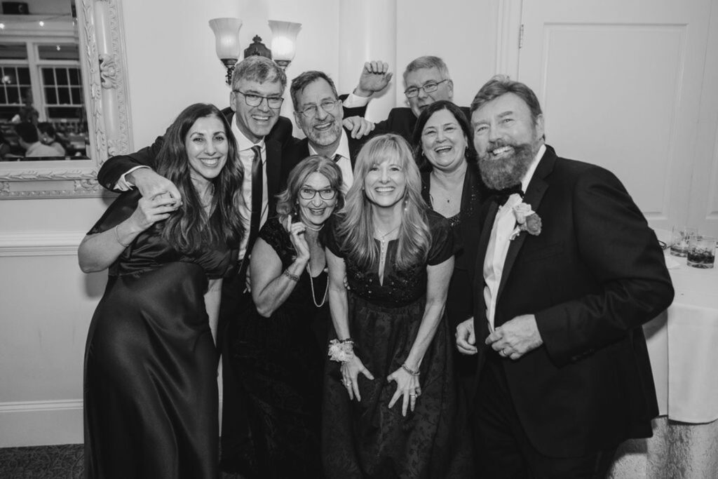 A group of eight adults dressed formally, smiling and posing together at a Chastain Horse Park wedding reception. The festive indoor setting features a table, drinks, and a mirror in the background, creating a joyful and celebratory mood.