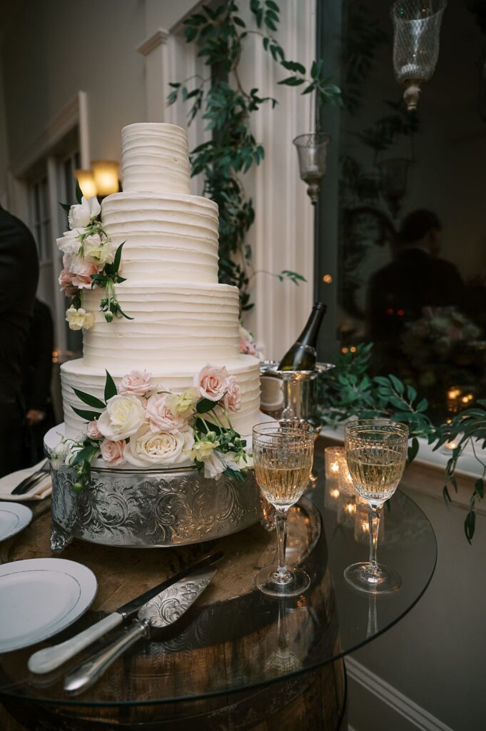A tall, white, tiered wedding cake decorated with pink and white flowers sits on a silver stand—perfect for a progressive buffet wedding in Atlanta. Two filled wine glasses, plates, and a cake knife rest nearby on a glass table.