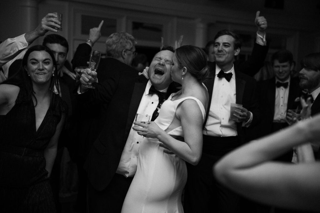 A group of people dressed formally celebrate at a progressive buffet wedding in Atlanta. In the center, a man laughs with his mouth open and holds a drink, while a woman in a light dress stands close to him, smiling as others cheer and raise glasses.