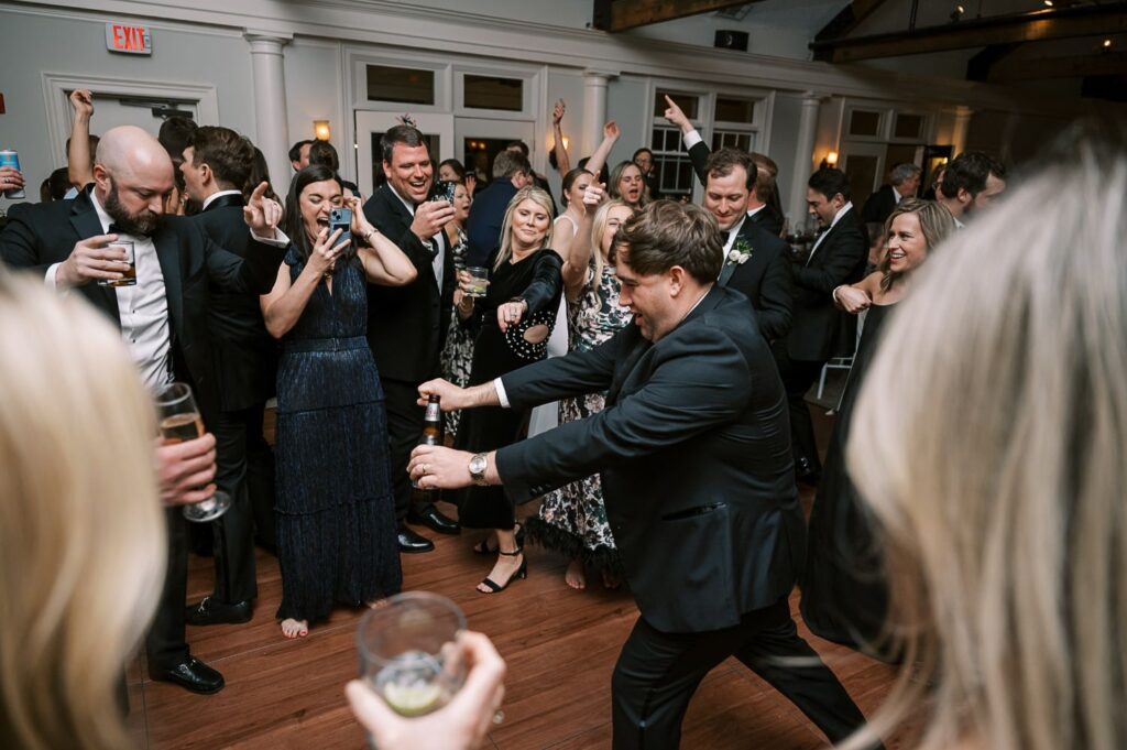 A group of people dressed formally are dancing and laughing at an indoor progressive buffet wedding in Atlanta, holding drinks and enjoying themselves on a wooden dance floor.