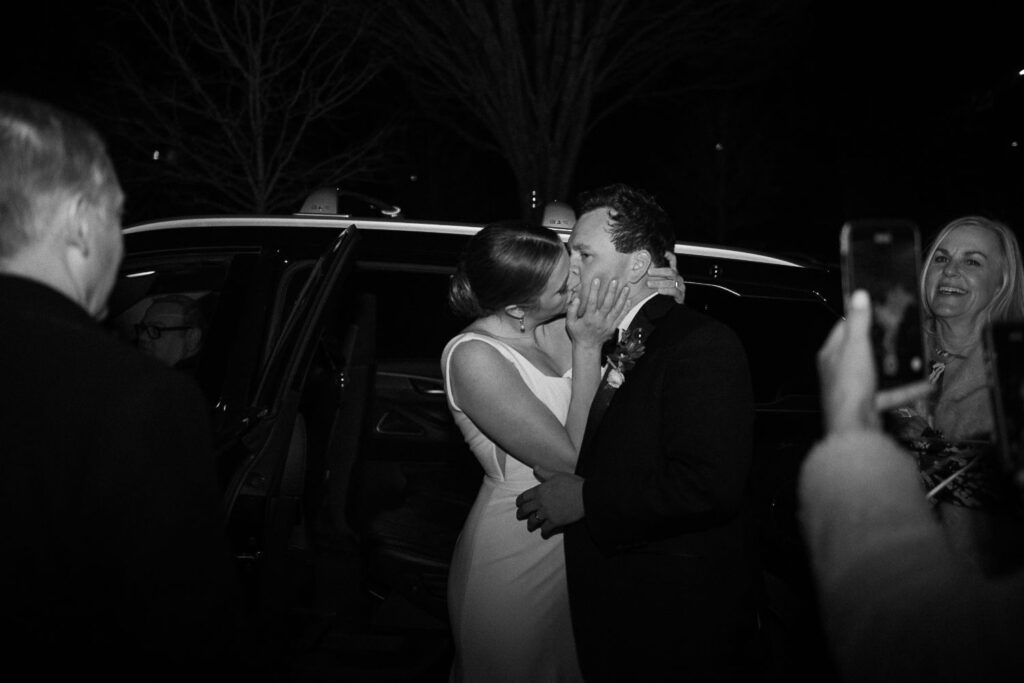 A bride and groom share a kiss at their progressive buffet wedding in Atlanta, surrounded by smiling guests capturing the joyful, celebratory moment on their phones in front of a car at night.