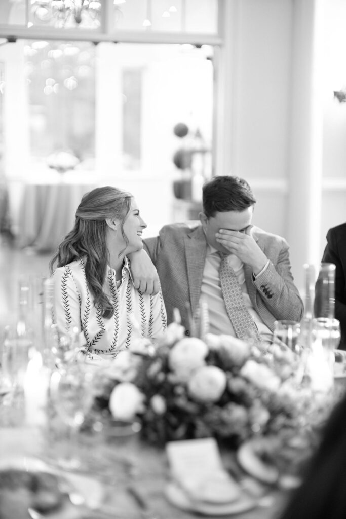 A woman and a man sit close together at a decorated table. The woman is smiling at the man, who has his arm around her and is covering his face with his hand. The setting is bright and elegant.