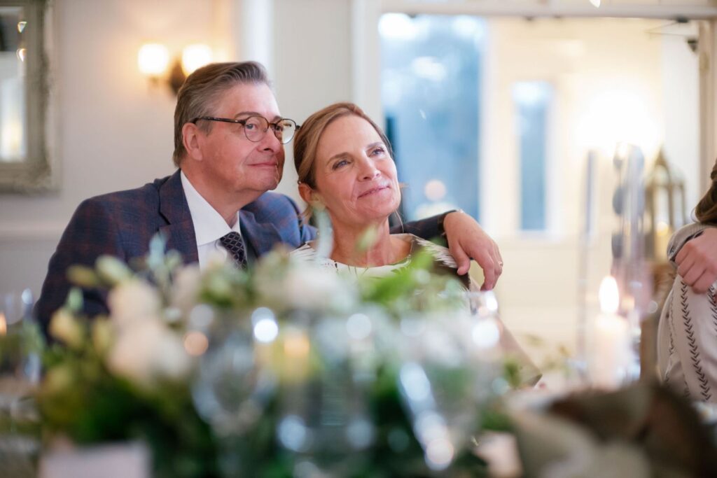 A middle-aged man and woman sit closely together at a decorated table, the man’s arm around the woman. Both look ahead and smile softly, surrounded by soft lighting and floral arrangements.
