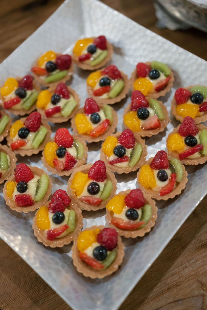 A tray of mini fruit tarts topped with a variety of fresh fruit slices, including strawberries, blueberries, kiwi, mandarin oranges, and raspberries, arranged neatly on a textured square platter.