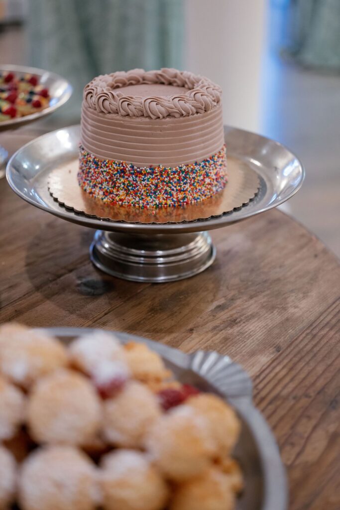 A chocolate frosted cake with colorful sprinkles along the sides sits on a silver cake stand atop a wooden table. Blurred pastries dusted with powdered sugar are visible in the foreground.