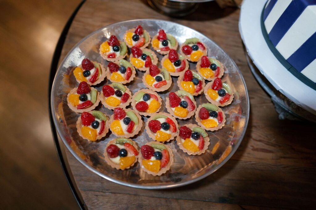 A round silver tray filled with small fruit tarts topped with slices of mandarin orange, berries, and kiwi, arranged on a wooden table next to a blue-and-white striped cake.