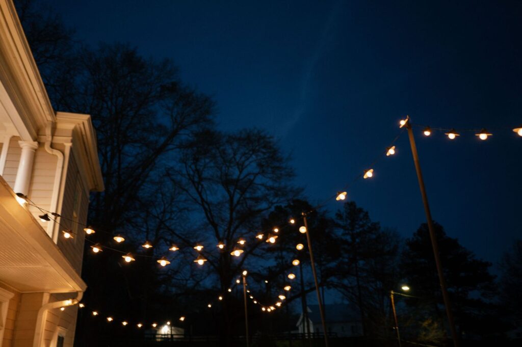String lights are hanging outdoors in the evening, glowing warmly against a deep blue sky. A house is visible on the left, and silhouettes of leafless trees can be seen in the background.
