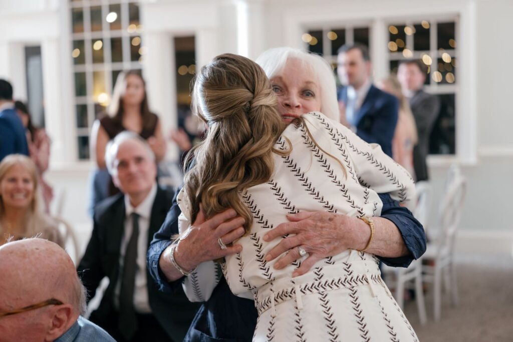 An elderly woman and a younger woman warmly hug at a formal indoor event, with seated guests and twinkling lights visible in the background.