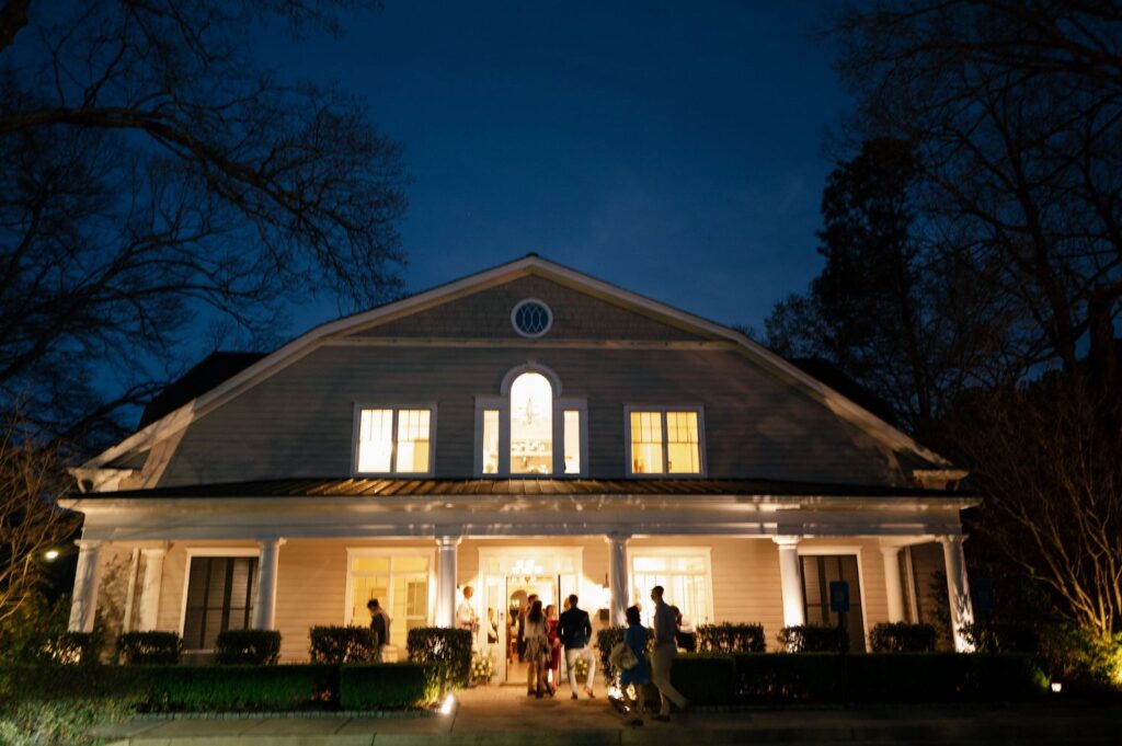 A large, well-lit house at night with people gathered near the front entrance, some standing under the porch, framed by leafless trees and a dark blue sky.