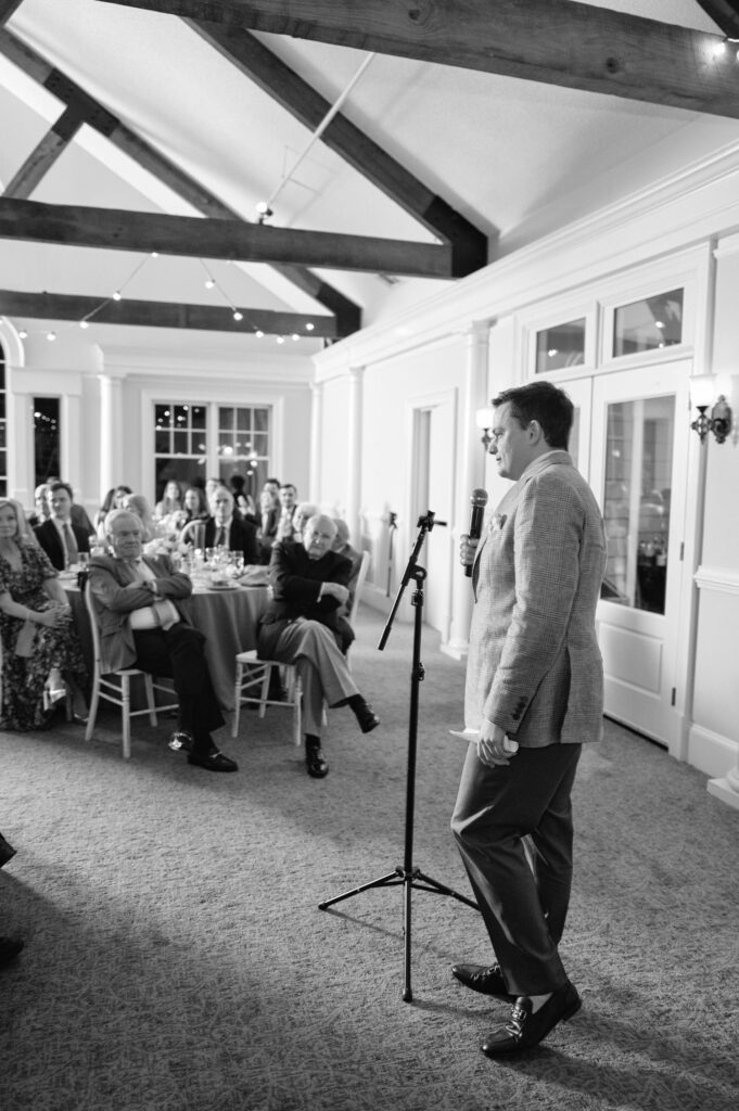 A man in a suit speaks into a microphone to a seated audience at a formal indoor event; string lights and wooden ceiling beams are visible above.