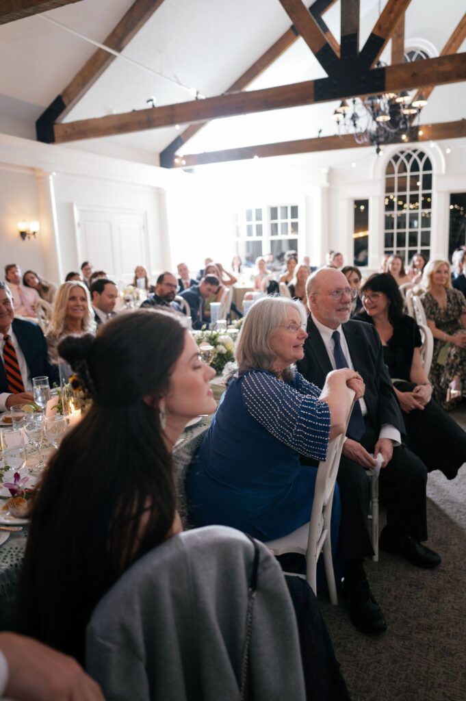 A group of people, dressed formally, sit at tables and smile while watching an event in a bright, airy room with exposed wooden beams and large windows.