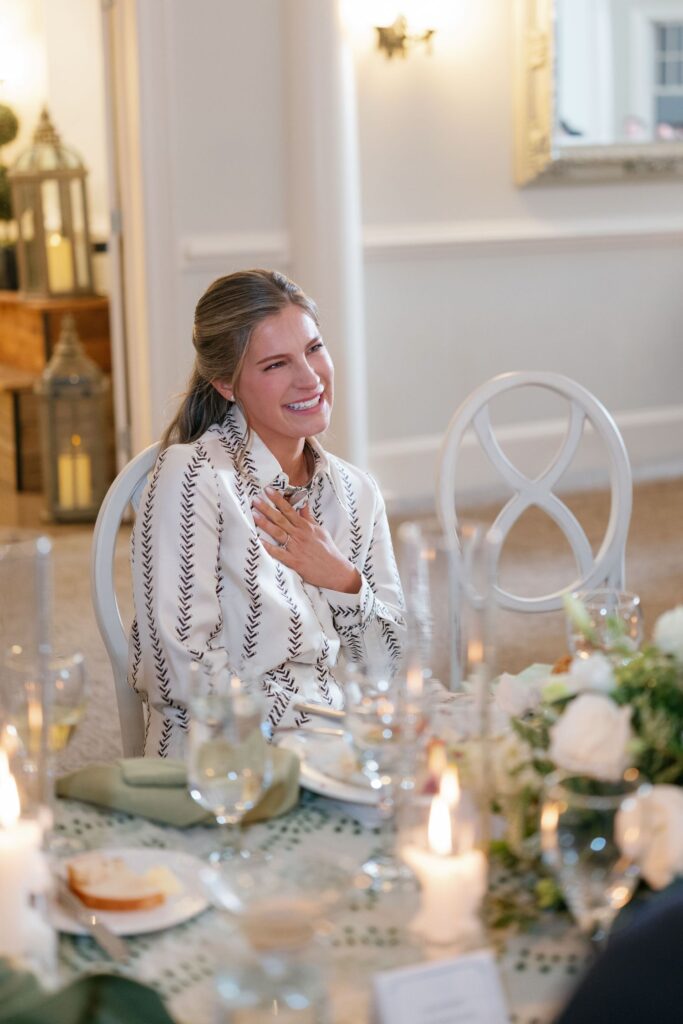A woman with long, light brown hair smiles and touches her chest while seated at a decorated dining table with candles, flowers, and glasses in a softly lit room.