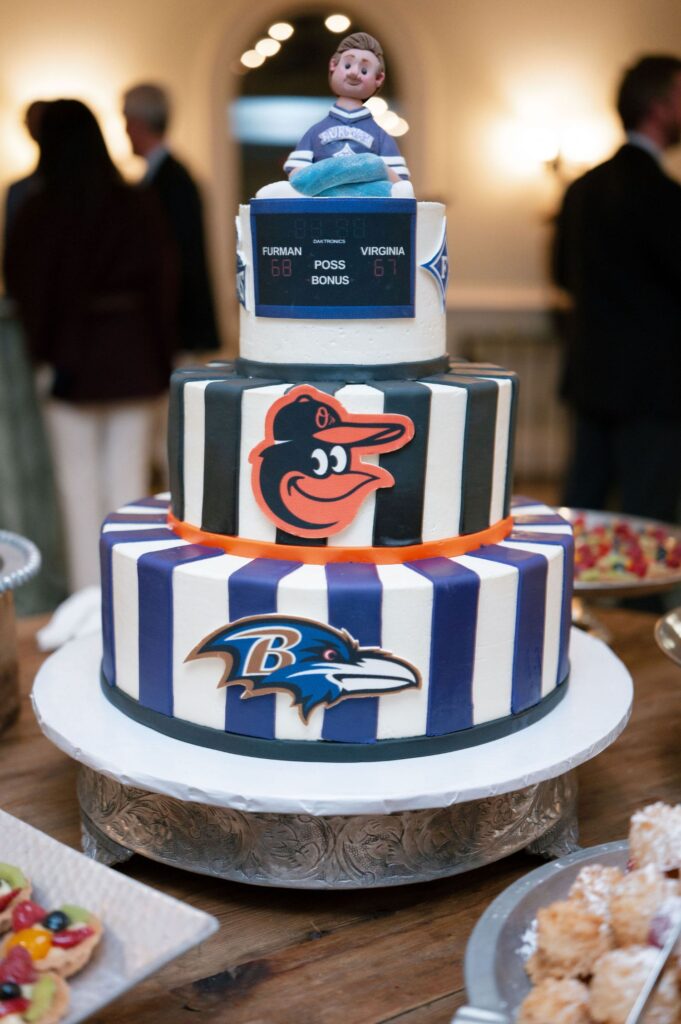 A three-tiered cake decorated with Baltimore Orioles and Ravens logos, purple and white stripes, and a scoreboard, topped with a figurine of a child in sports attire, sits on a table surrounded by desserts.