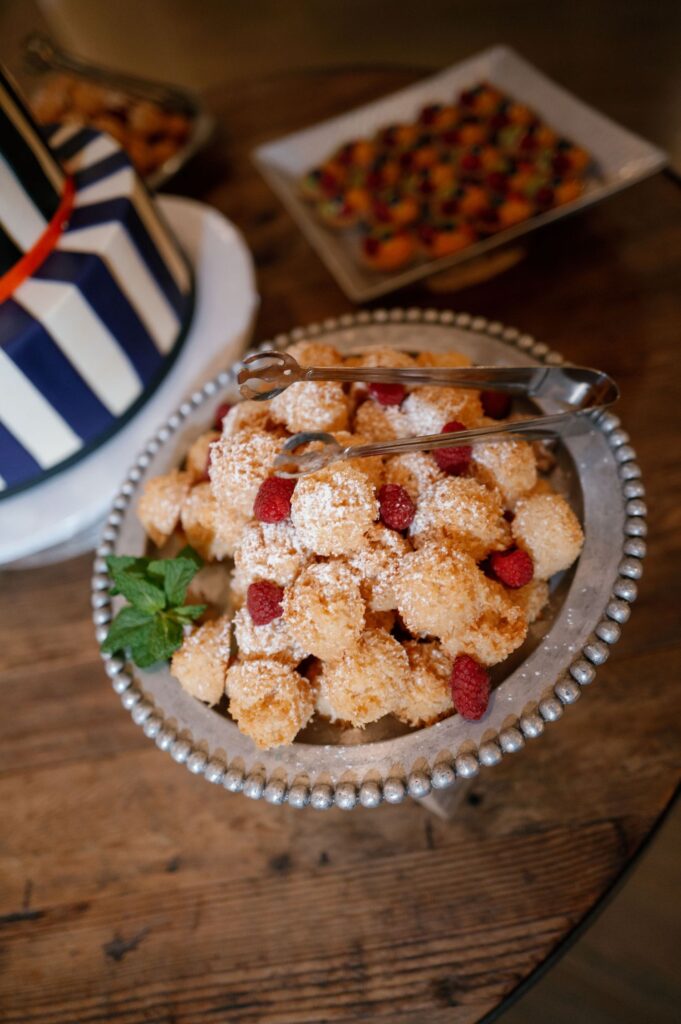 A silver bowl filled with powdered sugar-coated pastry bites and raspberries, garnished with a sprig of mint, sits on a wooden table next to a pair of tongs. Other desserts are visible in the background.