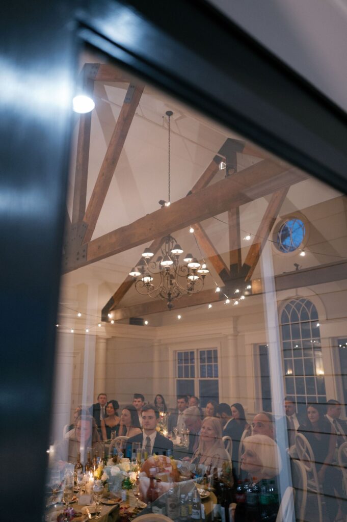 A group of people sit at decorated tables inside a well-lit room with wooden beams and a chandelier, seen through a window’s reflection.