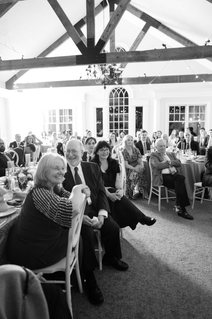 A group of people dressed formally sit at round tables in a bright, elegant venue with exposed wooden beams, smiling and enjoying an event. The mood is joyful and celebratory.