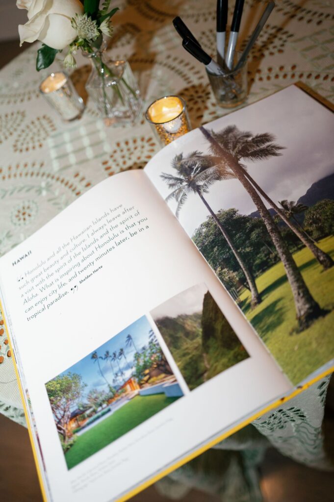 An open book on a lace-covered table displays photos of palm trees and lush scenery in Hawaii, alongside a paragraph of text. Nearby are candles, pens in a holder, and a vase with white roses.