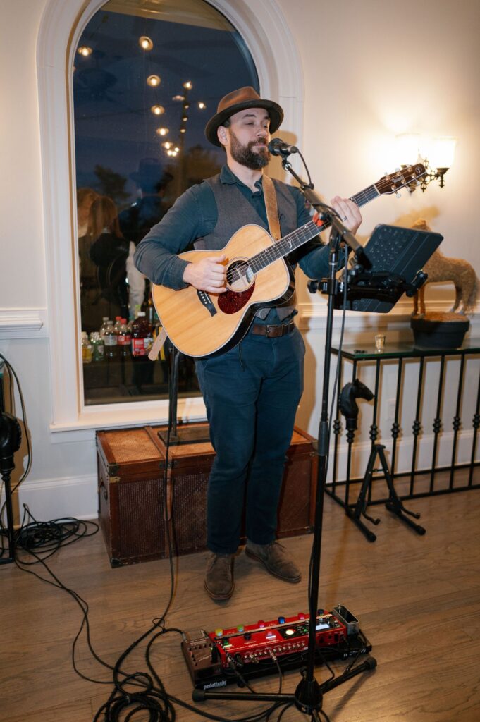 A man wearing a hat and vest plays an acoustic guitar and sings into a microphone indoors, with a pedalboard at his feet. A music stand, drinks, and a large window are visible in the background.