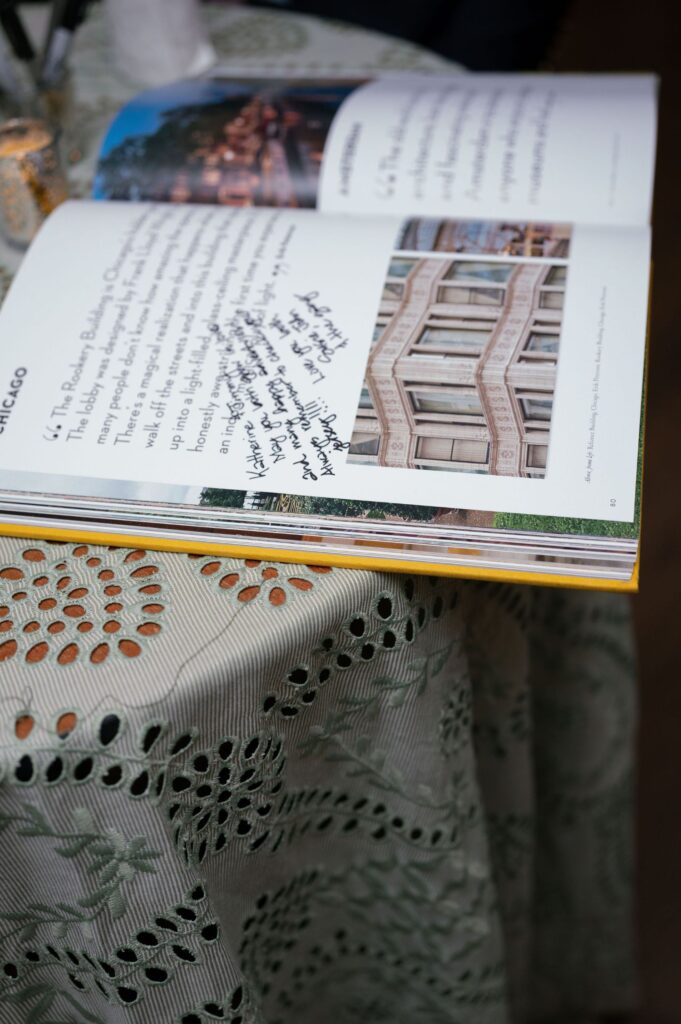 An open book with handwritten notes in black ink on its pages rests on a lace-patterned tablecloth. A building photo and printed text are visible in the book.