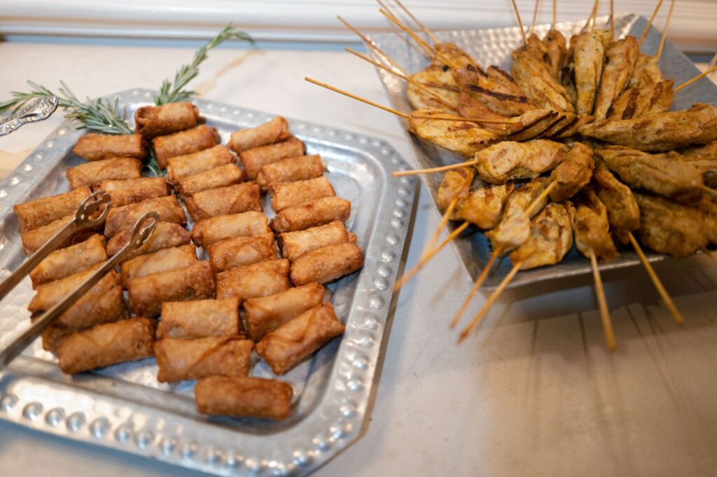 Two silver trays on a table, one filled with rows of golden-brown spring rolls and the other arranged with grilled chicken skewers. Metal tongs and a sprig of rosemary decorate the setup.
