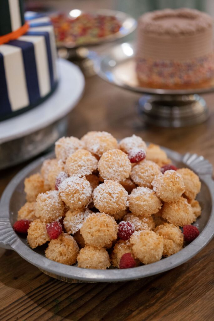 A metal tray filled with coconut macaroons dusted with powdered sugar and garnished with fresh raspberries, placed on a wooden table with cakes visible in the background.
