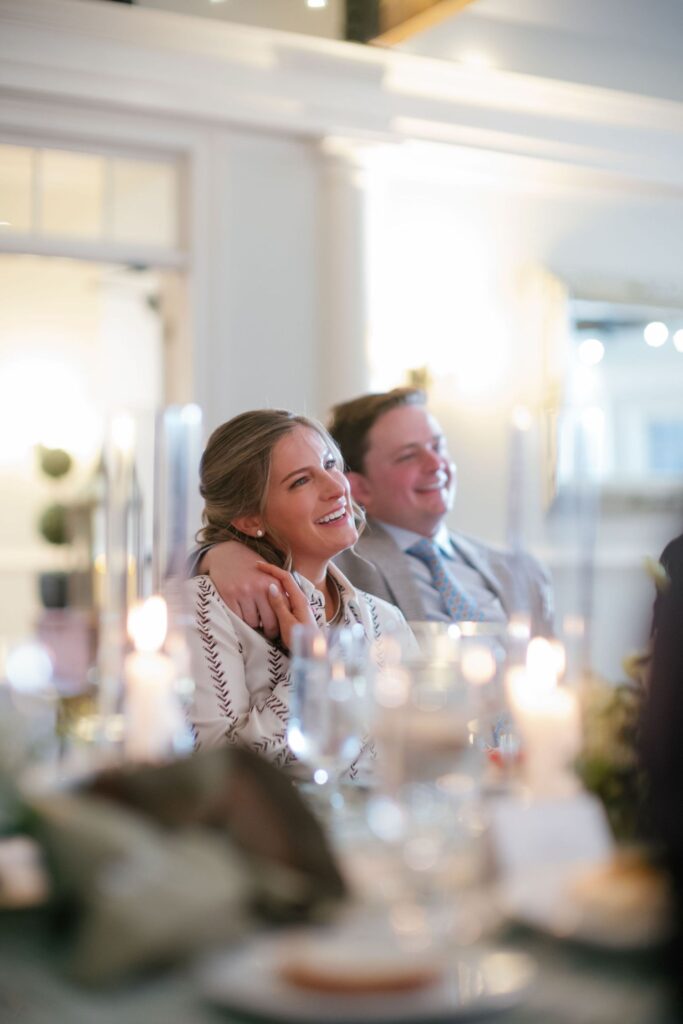 A smiling couple sits closely together at a candlelit table during a formal event, with the woman leaning back against the man, both looking happy and relaxed. The background is softly blurred.
