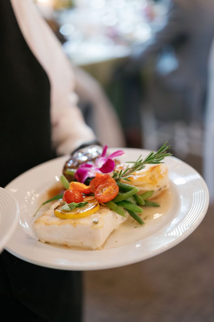 A white plate holds a fillet of fish topped with lemon slices, green beans, cherry tomatoes, a sprig of rosemary, and a purple orchid garnish. A person in white and black holds the plate.