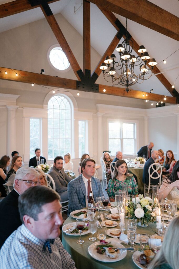 Guests seated at round tables enjoy a meal in an elegant, high-ceilinged venue with exposed wooden beams, arched windows, and a chandelier. Plates of food, drinks, and floral centerpieces are on the tables.