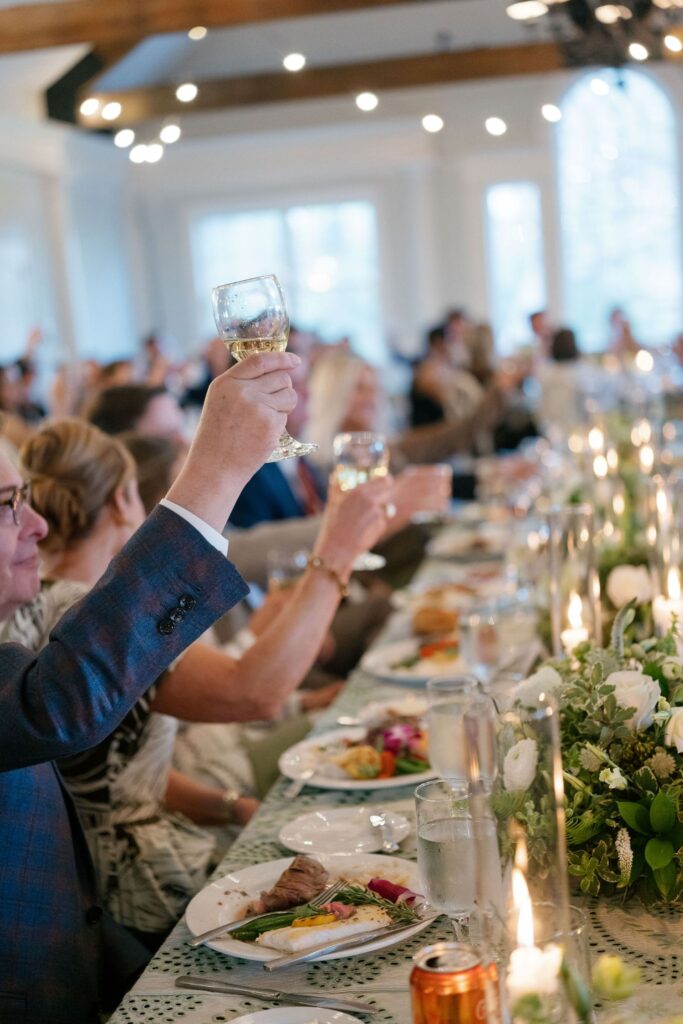 Guests seated at a long table raise glasses in a toast during a formal celebration, with plates of food, lit candles, and floral centerpieces decorating the table.