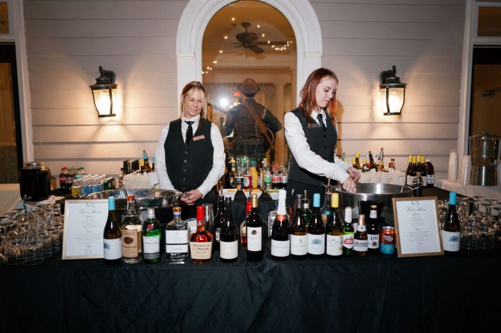 Two female bartenders in uniform stand behind a table filled with various liquor bottles, mixers, and glasses. The setting appears to be an outdoor event, with a lit house wall and lanterns in the background.