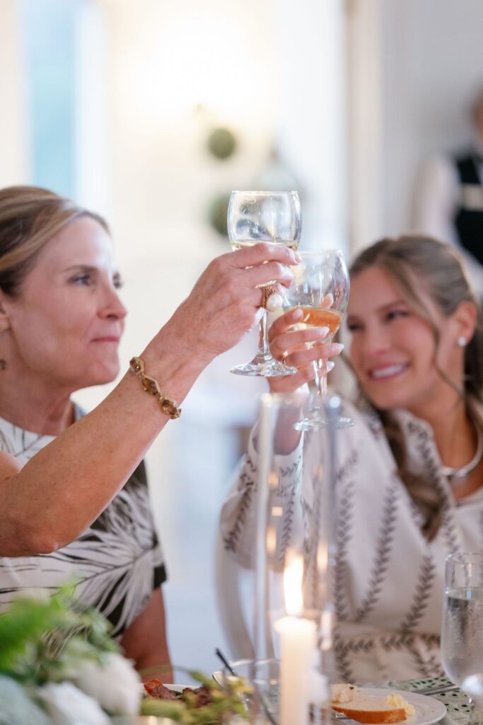 Two women smiling and raising wine glasses in a toast at a well-lit, elegant dining setting, with plates of food and candles visible on the table in the foreground.