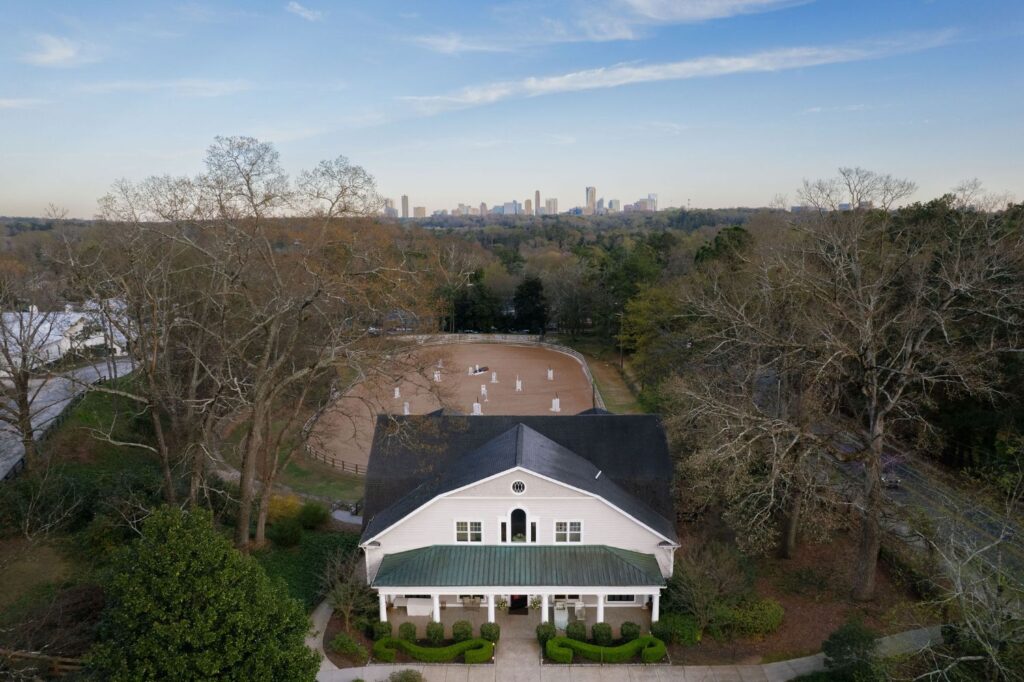 Aerial view of a white building with a green roof in front of a sandy equestrian arena, surrounded by trees, with a city skyline visible in the distant background under a blue sky.