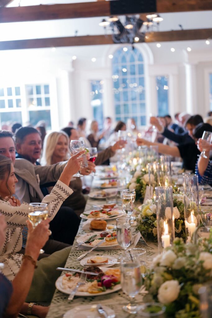 A group of people sitting at a long table raise their glasses in a toast during a formal meal. The table is elegantly decorated with flowers, candles, and plates of food. The atmosphere is bright and festive.