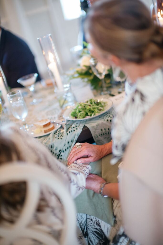 Two women sit at a decorated dining table, one resting her hand gently on the others. A plate of salad, bread, candles, and glassware are visible. The scene feels warm and intimate, suggesting comfort and support.