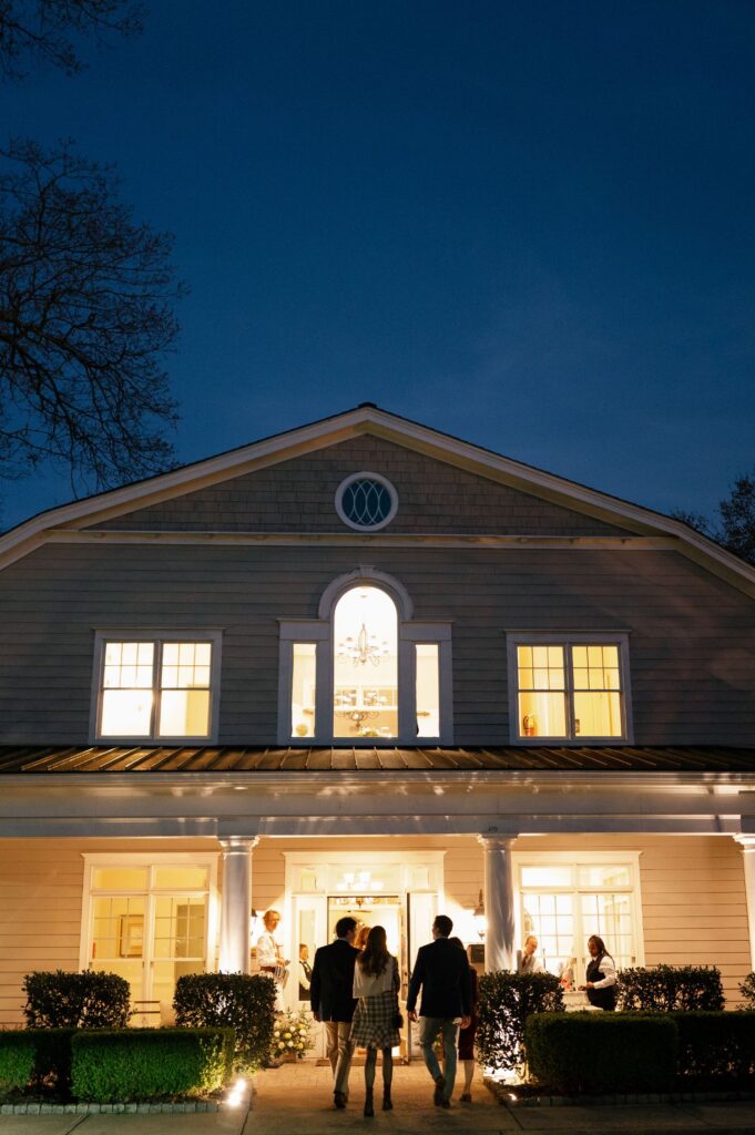 People walk toward a brightly lit house with white columns at dusk. The windows glow warmly, and a few people are visible inside, creating a welcoming atmosphere against the darkening sky.
