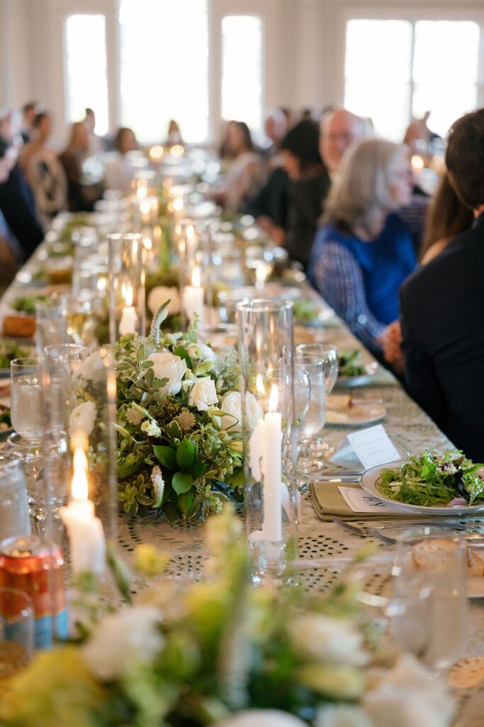 A long banquet table is set with floral centerpieces, lit candles, plates of salad, and glassware. Guests are seated on both sides, engaged in conversation in a bright, elegant room.
