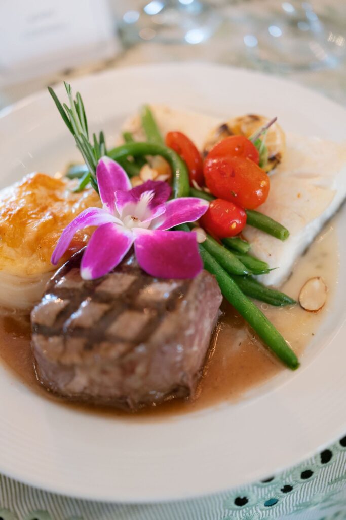 A plated gourmet meal features grilled steak, a white fish fillet, green beans, cherry tomatoes, a potato gratin, and a purple orchid garnish on a white dish.