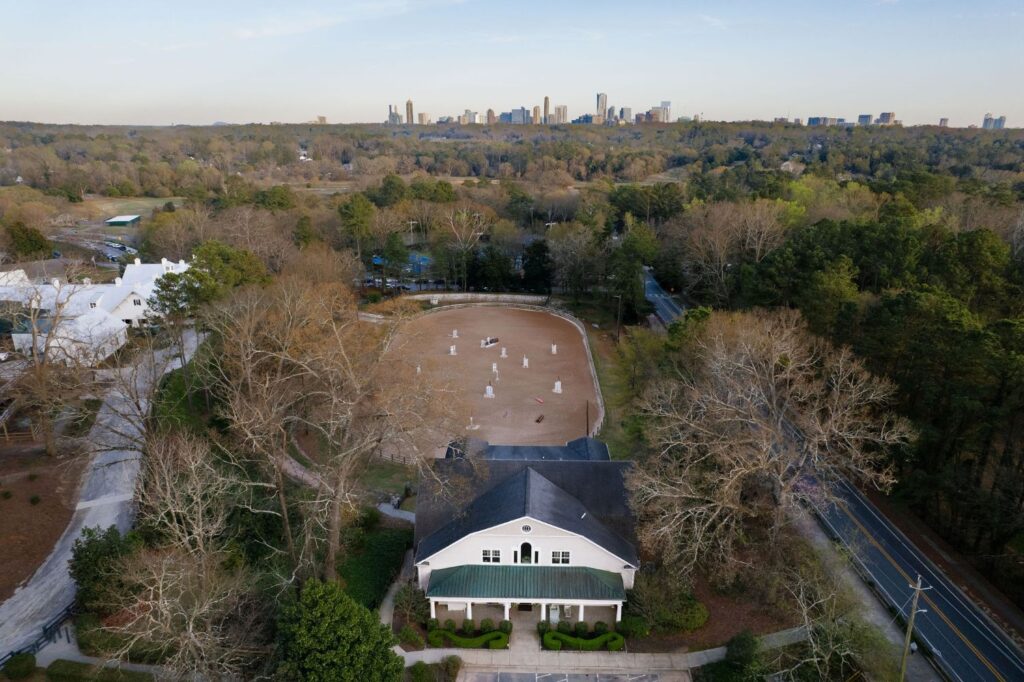 Aerial view of a large white clubhouse in front of a sandy equestrian arena with jumps, surrounded by trees. In the distance, a city skyline rises above the forested landscape.