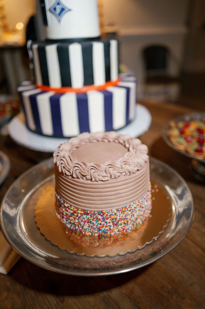 A small chocolate cake with piped frosting and colorful sprinkles on the sides sits on a silver platter; a larger striped cake is blurred in the background.
