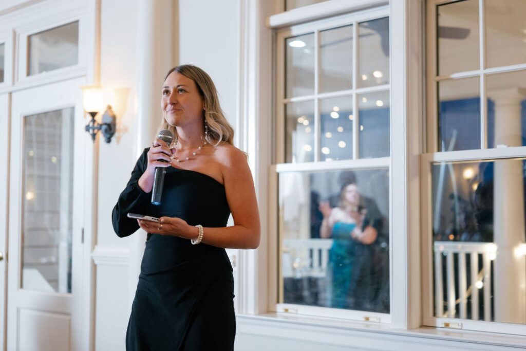 A woman in a black, one-shoulder dress holds a microphone and phone, speaking indoors. There are large windows behind her, with lights and a reflection of another person visible outside.